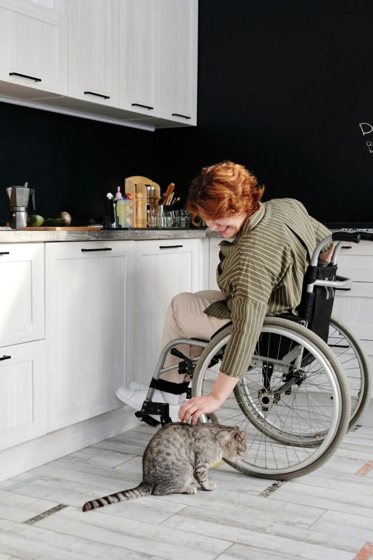 A woman in a wheelchair smiles while petting a tabby cat in a modern kitchen.